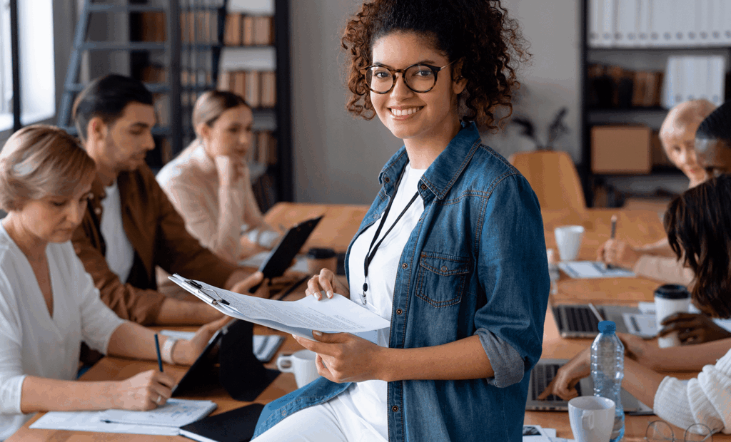 DEI. Imagem de uma biblioteca com algumas pessoas estudando e no centro da imagem em foco temos uma menina negra de pele pouco retinta, usando óculos e sorrindo, segurando uma prancheta. 