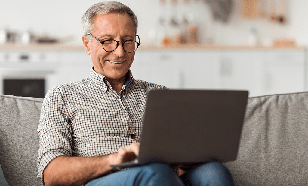 Acessível. Imagem de um senhor de idade de cabelos grisalhos sorrindo e usando óculos, ele está sentado no sofá com um notebook em sua perna.